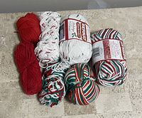 Seven skeins of dishcloth yarn in holiday colors and patterns arranged on a floor surface, including solid red, white with red and green flecks, and striped patterns in red, green, and white.