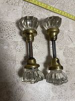 Two vintage style glass door handles with brass fixtures laid on tiled surface with tape measure for scale about 12 inches long.