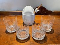 Front view showing five crystal old fashioned glasses and white ceramic honey pot with lid, honey dipper and masher on top, all placed on wooden furniture.