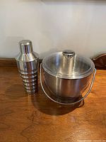 Photo showing both the stainless steel ice bucket with lid and handle, and the cocktail shaker beside it on a wooden surface.