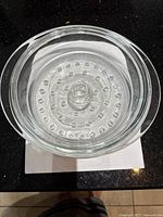 Top view of a clear round glass steamer insert with small raised bumps inside and central handle.