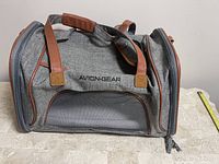 Front and side view of grey soft shell cat carrier with brown trim, showing the Avion-Gear brand name and mesh ventilation panels.