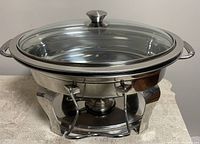 Front view of stainless steel oval chafing dish with glass lid and fuel holder on a table.