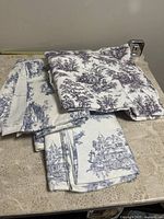 Various folded pieces of upholstery fabric showing white background with scenic house patterns in blue and purple tones.