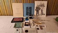 View of all items arranged on a white surface, showing the art prints, tie clip, nail file, silver spoon, small containers, and assorted collectibles.