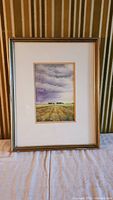 Framed watercolor painting of a rural landscape with field and cloudy sky, shown from a distance, including the entire frame.