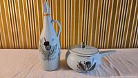 Tall stoneware decanter with painted thistle design and stopper, next to lidded pot with handle and similar decoration