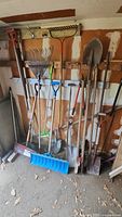 Photo showing various garden tools including rakes, shovels, snow pusher, and hoes hung on a pegboard in a garage space with a wall behind them.