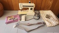 Kenmore 158A sewing machine with foot pedal, spool of thread, metal clamp, beige quilting pad, and Dexter Hemmer & Stitcher accessory box on table.