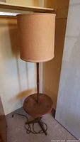 Full view of lamp showing tan fabric drum shade, teak column, round side table, and tripod base