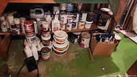 Wide view of underside of workbench showing all paint cans and containers grouped in three main clusters on green floor including some plastic bottles and boxes.
