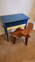 Desk with blue top and legs, yellow drawer, and matching knobs; wear and paint chipping visible; positioned beside wooden curved 3-legged chair