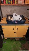 Photo of wooden rolling workshop cart with yellow cabinet doors. A blue plastic bin on top contains a white plastic bag and electrical parts.