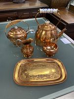 Five-piece antique copper tea set arranged on a glass surface, showing the large copper teapot with ceramic handle, copper teapot with wicker handle, creamer, bowl, and engraved tray.