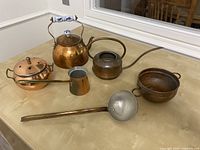 Full lot picture showing five copper and brass items on a table: teapot, watering can, potpourri container, small cup, and ladle.