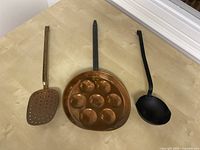 Top view of the three items laid flat on a wooden surface; copper skimmer on left, copper aebleskiver pan in center, cast iron ladle on right