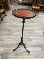 View of the antique wood pedestal side table showing the round tray top with red leather inset and tripod base on light colored floor with other furniture in background.