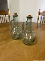 Two clear glass decanters with handles and cork stoppers set on a wooden table with chairs in background