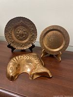 Front view of the set of three decorative plates, two round with embossed ethnic designs and one fish-shaped plate, showing brass/copper metal finish and detail.