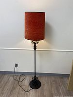 Full view of the floral patterned floor lamp turned off, showing cylindrical reddish-brown fabric shade and ornate metal base and stand with cord on hardwood floor against a light wall.