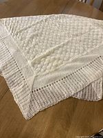 Photo showing the folded cream wool baby blanket with textured knit and airy mesh border on wood table.