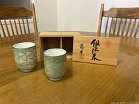 Two porcelain tea cups with green bamboo pattern placed next to wooden box with Japanese script on lid.