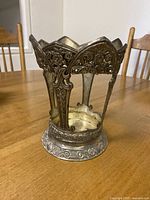 Front angled view of the ornate silver vase holder showing detailed floral and scroll design on metal panels and scalloped top edge.