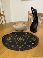 Three-item set featuring hand-painted floral wood tray, beige ceramic bowl with textured inside, and smooth black wooden abstract sculpture on a wooden table.