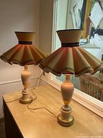Pair of vintage ceramic table lamps featuring beige bases with gold accents and beige fabric lampshades, one flared with scalloped trim and one drum-shaped, displayed on a wooden table by window.