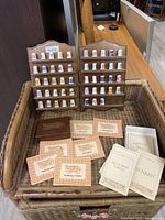 Two wooden display racks holding 48 porcelain thimbles with various colorful designs, arranged in rows. Several themed informational cards are placed in front of the racks inside a wicker basket.