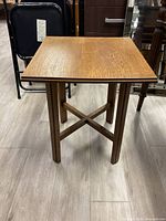 Full front view of rustic light brown wooden side table showing square top, four straight legs and X-shaped supports on a light floor.