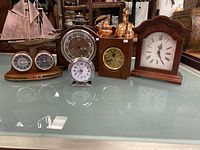 Set of five mantel clocks on glass surface, showing styles from nautical to vintage, including a wooden nautical clock set with miniature sailboat and instruments, a round metal-faced clock, a silver wind-up alarm clock, a wooden rectangular clock with brass face, and an arch-top wooden mantel clock.