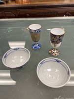 Two blue and white floral porcelain bowls and two multicolored floral goblet-style cups with gold accents on glass table.