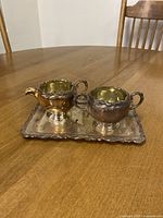 Full view of the brass sugar bowl and creamer placed on the rectangular brass tray on wooden table, showing ornate floral patterns and reflective tarnished surfaces.
