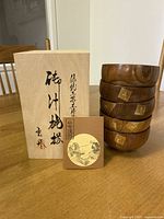 Five stacked lacquered wood bowls with natural wood exterior and gold accents. Wooden box with Japanese calligraphy and branding card on table.