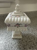 Side angle showing the pedestal base, scalloped edge lid, and floral hand-painted decoration on the milk glass box.