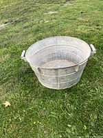 Front angle view of antique metal washtub sitting on grass, showing handles and circular ribbed interior base.