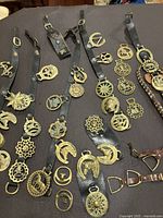 Overview of several black leather horse harness straps laid out, each decorated with multiple ornate brass medallions featuring horseshoes, star shapes, and other classical symbols, alongside a small leather whip and metal stirrups.