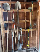 Photo showing various gardening tools hanging on a wooden wall including 3 shovels, 2 rakes, trimming shears, and additional hand tools.