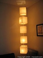 Lit view of hanging light showing five illuminated square fabric shades hanging vertically from ceiling.