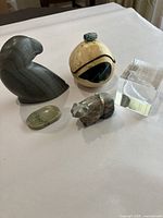 Four stone carvings and paperweights on white surface including gray bird carving, small animal figure with beads, green stone oval, and indigenous painted round piece.