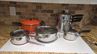 Photo of all pots and steamers arranged on white surface on kitchen counter, showing one orange pot and multiple stainless steel sauce pans and steamers with black handles and glass lids.