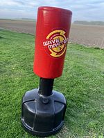 Front angled view of red Wavemaster punching bag on black resin plastic base sitting on grass outside in daylight against a field background.