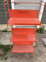 Front view of orange metal shelving unit showing three shelves and top rail detail. Visible surface dirt and wear.