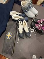 Vintage ski patrol rescue bag alongside four pairs of ski boots in various sizes and colors arranged on a dark surface.