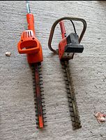 Top view of two electric hedge trimmers side by side on concrete floor, showing blades and handles.