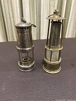 Two vintage coal mining safety lamps placed on a dark surface, one brass-colored and one silver-colored, photographed against a curtain background.