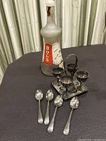 Photo showing the vintage Bols liquor bottle, frosted glass with original red and white label, four silver plated egg cups arranged on holder, and five matching spoons on a dark surface.