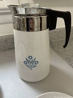 Side view of the vintage Corning stove top percolator showing white body with blue floral pattern, metal top, and black handle