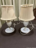 Pair of vintage hobnail milk glass lamps shown together on a table with cords visible and white fabric shades trimmed with pom-pom edging.
